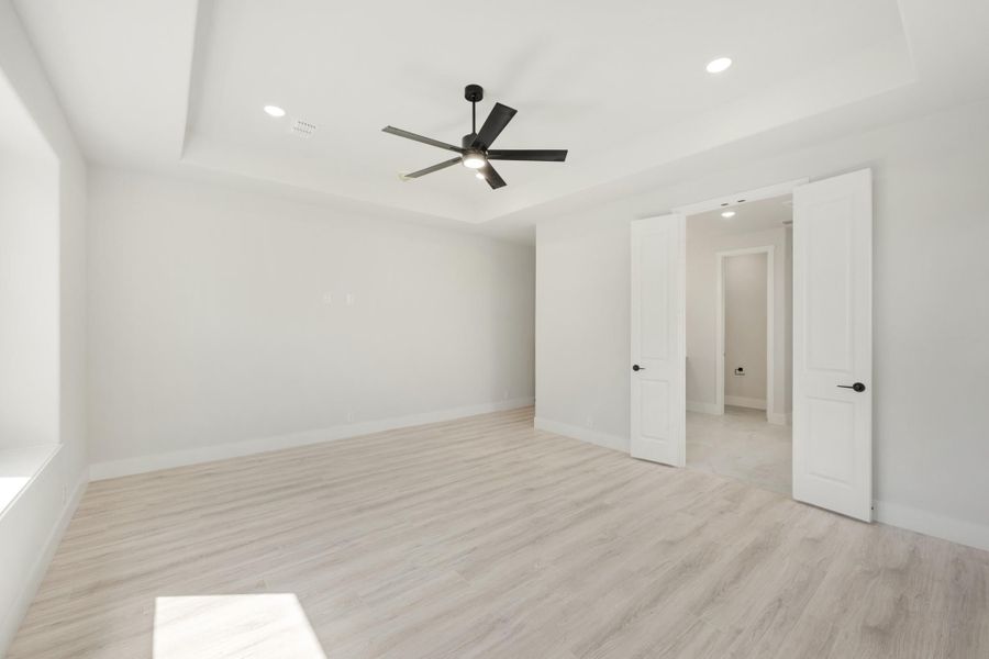 Empty bedroom with tray ceiling, black ceiling fan, light wood floors, and double doors leading to hallway