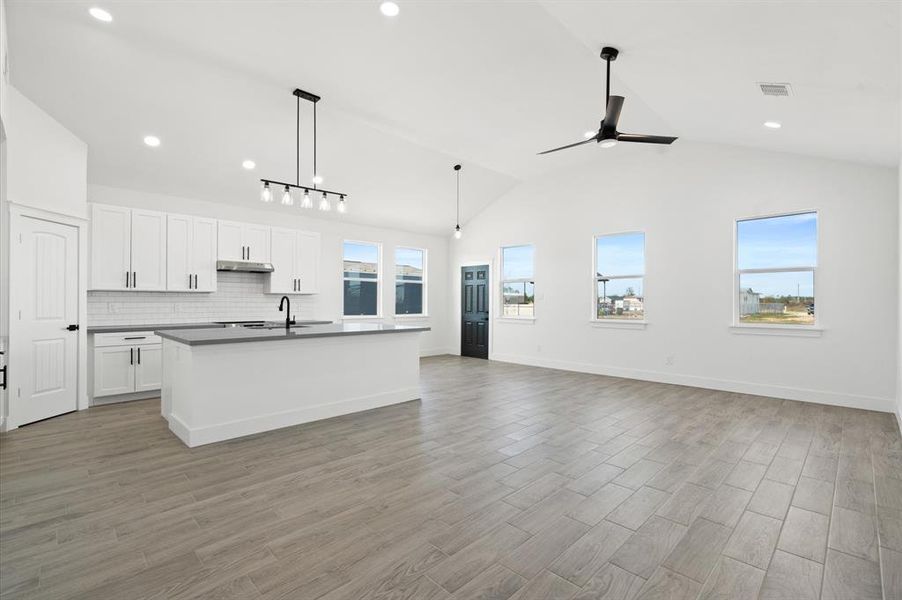 Modern, open-concept kitchen and living space with high ceilings, sleek white cabinetry, a central island with a sink, and contemporary lighting. Large windows provide ample natural light.