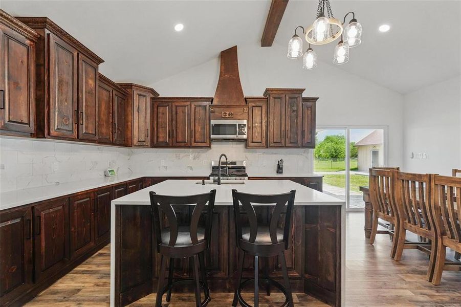 Kitchen with backsplash, dark brown cabinets, a center island with sink, light wood finished floors, and beamed ceiling Kitchen with backsplash, dark brown cabinets, a center island with sink, light wood finished floors, and beamed ceiling