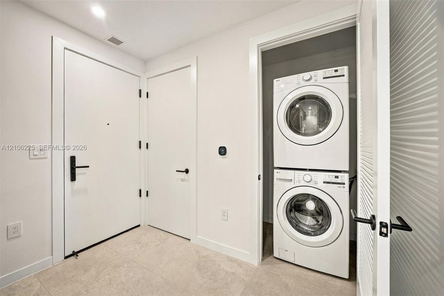 Laundry area featuring a stacked washer and dryer, neutral tile flooring, white slab doors, and matte black hardware Laundry area featuring a stacked washer and dryer, neutral tile flooring, white slab doors, and matte black hardware