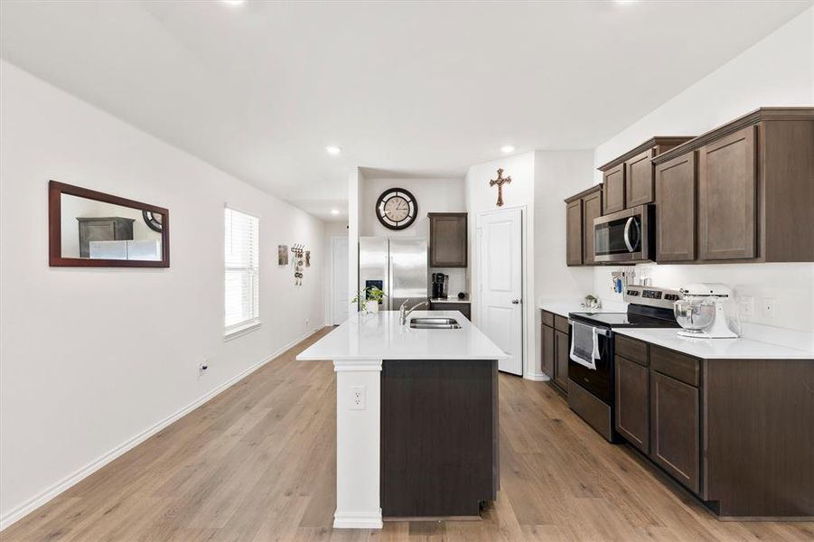 Kitchen with stainless steel appliances, dark wood finish cabinetry, a center island with sink, light wood finished floors, and light stone counters Kitchen with stainless steel appliances, dark wood finish cabinetry, a center island with sink, light wood finished floors, and light stone counters