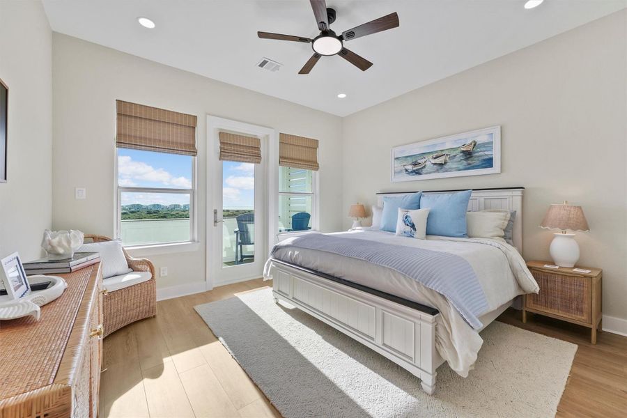 The third bedroom is located on the second floor and could be used as a second primary suite. It features furnishings from Pottery Barn and Serena & Lily such as a King bed, and matching dresser and nightstands and a framed Samsung TV. Large windows and a glass hurricane rated door leading to a balcony with scenic views. It has a cozy, coastal decor with light  flooring, a ceiling fan, wicker accents, and a plush bed adorned with soft blue and white linens. Ensuite bathroom and walk in closet.
