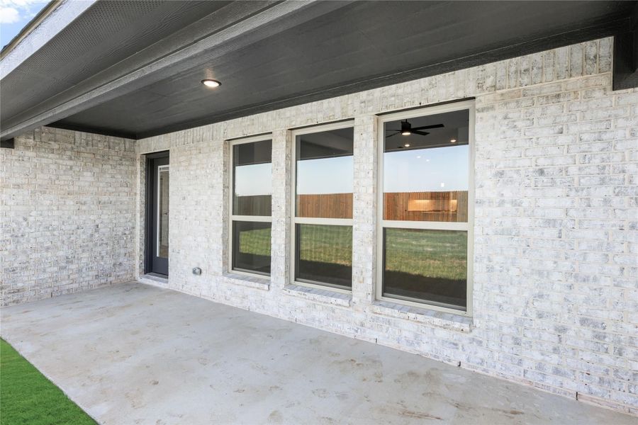 Covered concrete patio with light brick exterior, three large windows, and a dark entry door