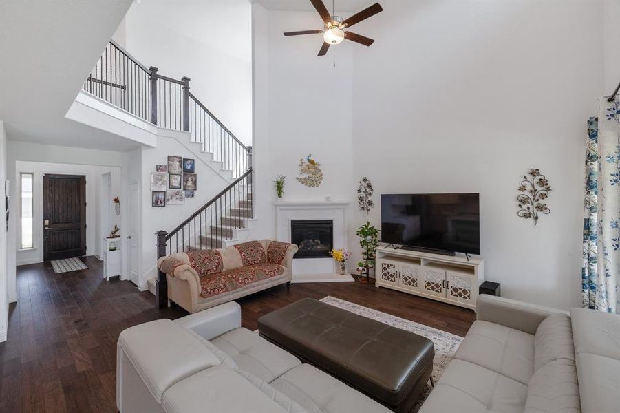 Living room featuring dark wood finished floors, a ceiling fan, a glass covered fireplace, a towering ceiling, and stairs