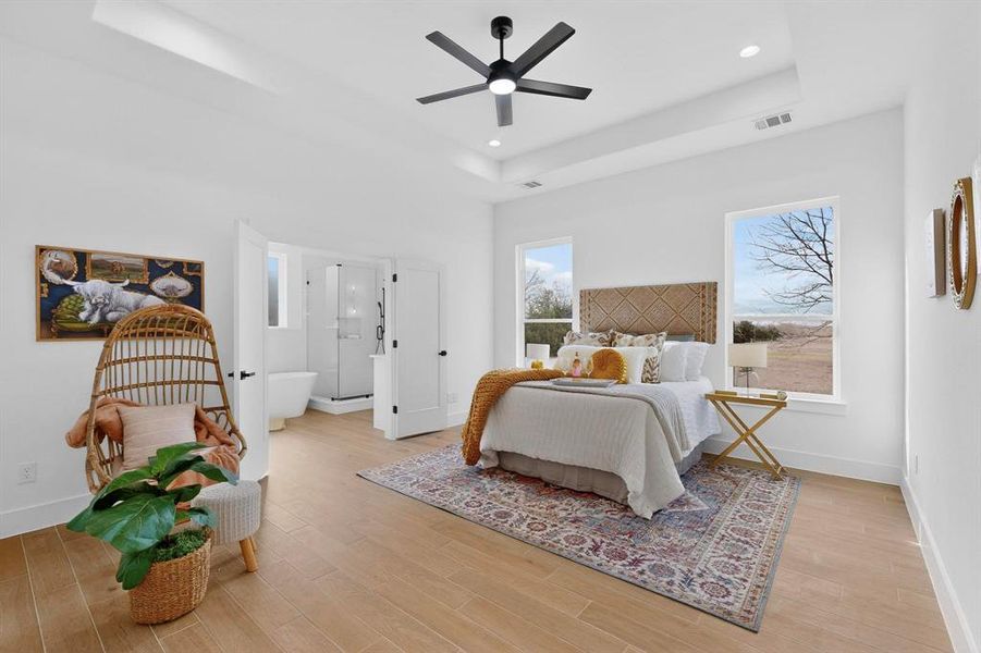 Bedroom with light wood-style flooring, a ceiling fan, ensuite bathroom, a raised ceiling, and recessed lighting