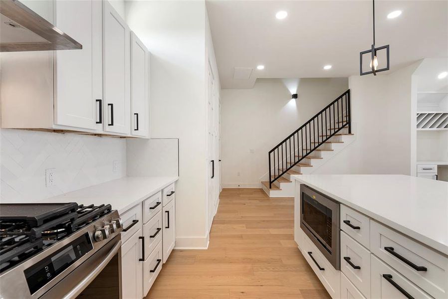 This is a modern kitchen with white cabinetry, stainless steel appliances, and herringbone backsplash, complemented by natural wood flooring. The space opens up to a staircase with a sleek black railing, leading to the upper level.