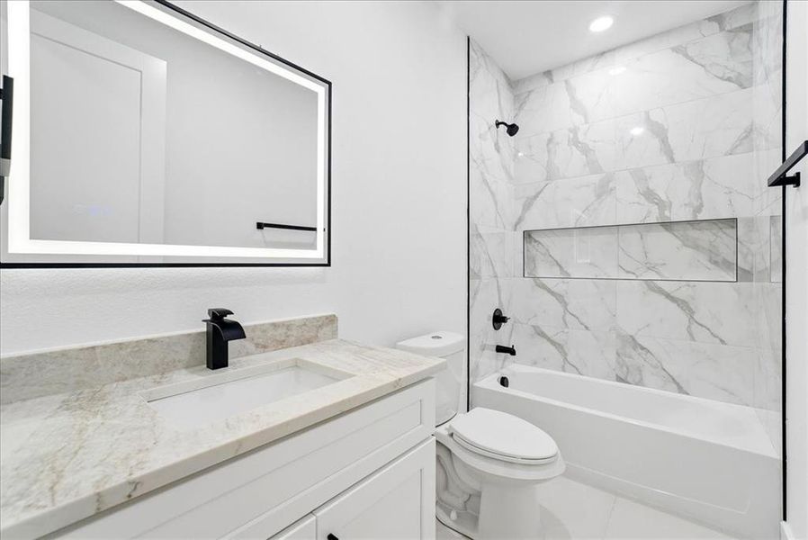 Bathroom featuring a lighted vanity mirror, waterfall-style faucet, light-toned countertop, and a shower/tub combination with a tiled surround Bathroom featuring a lighted vanity mirror, waterfall-style faucet, light-toned countertop, and a shower/tub combination with a tiled surround