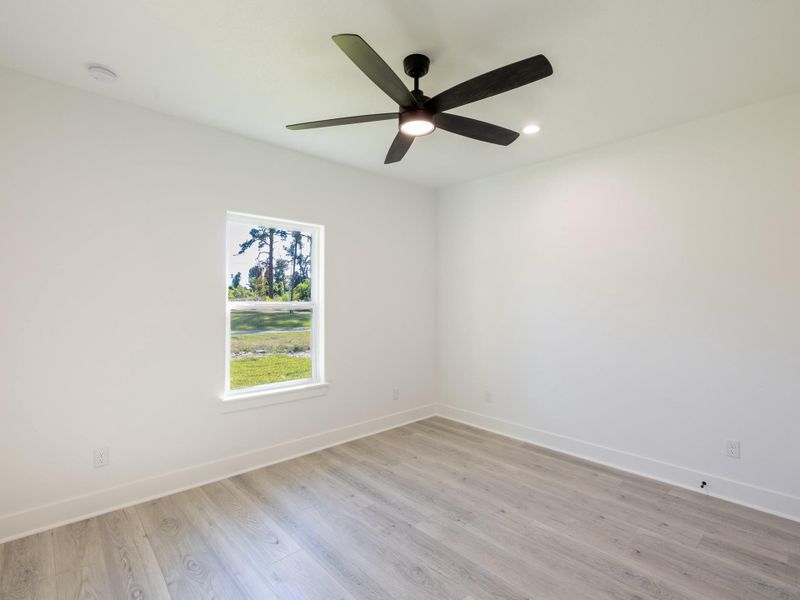This room features bright, neutral walls and light wood flooring, creating an airy feel. A large window provides ample natural light and offers a view of the outdoors. The ceiling is equipped with a modern black fan and recessed lighting.