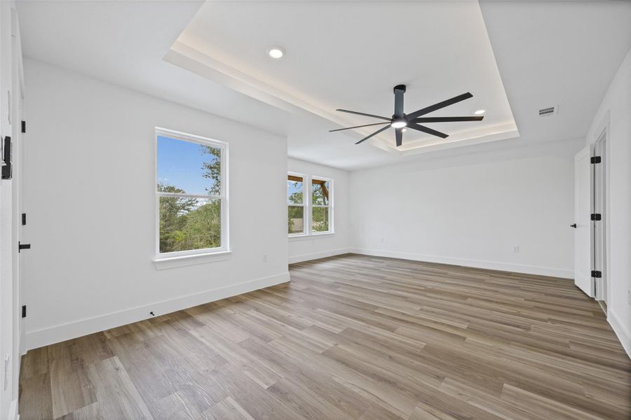 Spacious primary bedroom featuring wood-finish flooring, a tray ceiling with recessed lighting, and a modern ceiling fan Spacious primary bedroom featuring wood-finish flooring, a tray ceiling with recessed lighting, and a modern ceiling fan