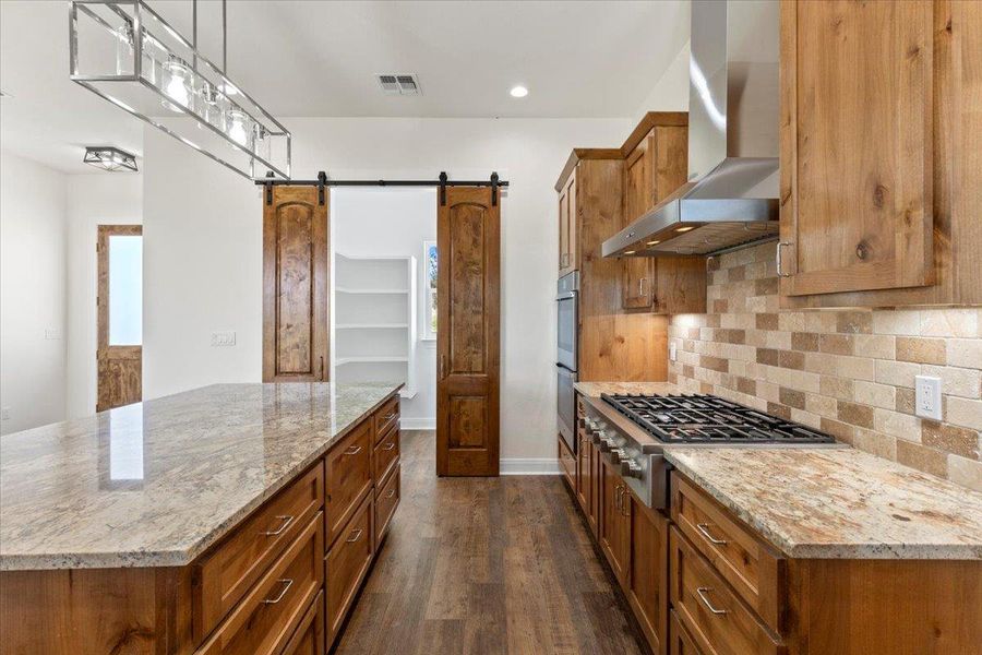 Kitchen with wall chimney range hood, appliances with stainless steel finishes, a barn door, brown cabinets, and dark wood-style floors