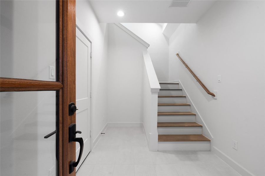 The front entry opens to a clean, minimalist foyer with light tile flooring, a wood-accented staircase, and access to the first-floor bedroom suite. The space establishes the home’s neutral palette and modern finishes.
