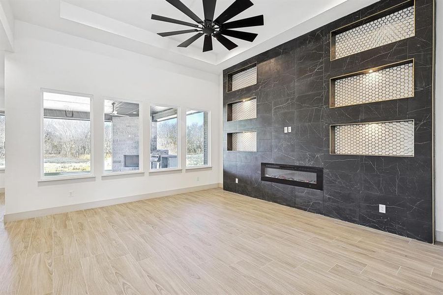 Unfurnished living room featuring light wood-type flooring, a ceiling fan, a tray ceiling, and a tiled fireplace Unfurnished living room featuring light wood-type flooring, a ceiling fan, a tray ceiling, and a tiled fireplace