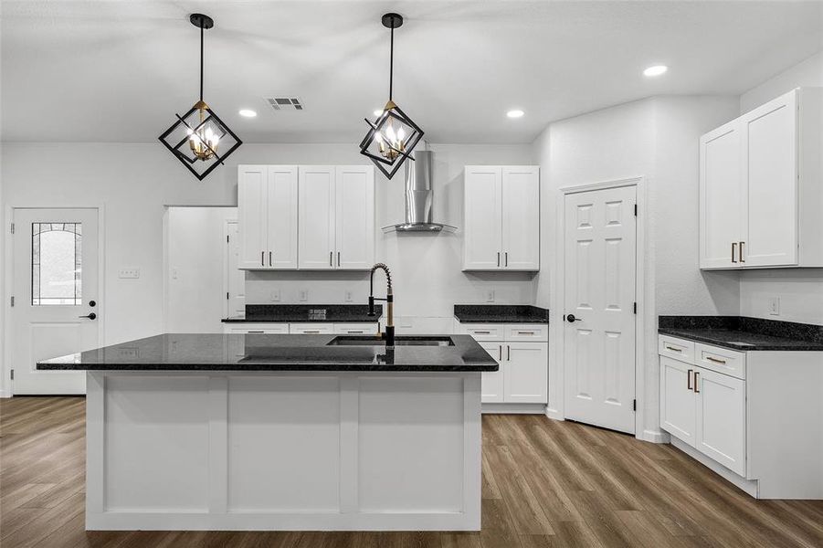 Contemporary kitchen featuring a large central island with a sink and dark countertop, white shaker-style cabinetry, stainless steel range hood, recessed lighting, and wood-finish flooring Contemporary kitchen featuring a large central island with a sink and dark countertop, white shaker-style cabinetry, stainless steel range hood, recessed lighting, and wood-finish flooring