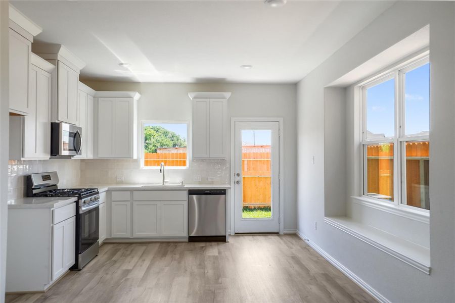 The kitchen features large windows, access to the backyard, stainless steel appliances, white cabinets, and tile backsplash.