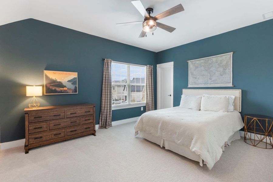 This bedroom features a cozy layout with a blue accent wall, a comfortable bed, and a ceiling fan. Natural light streams in through a window with plaid curtains. There's a wooden dresser and decorative wall art, creating a warm and inviting atmosphere.