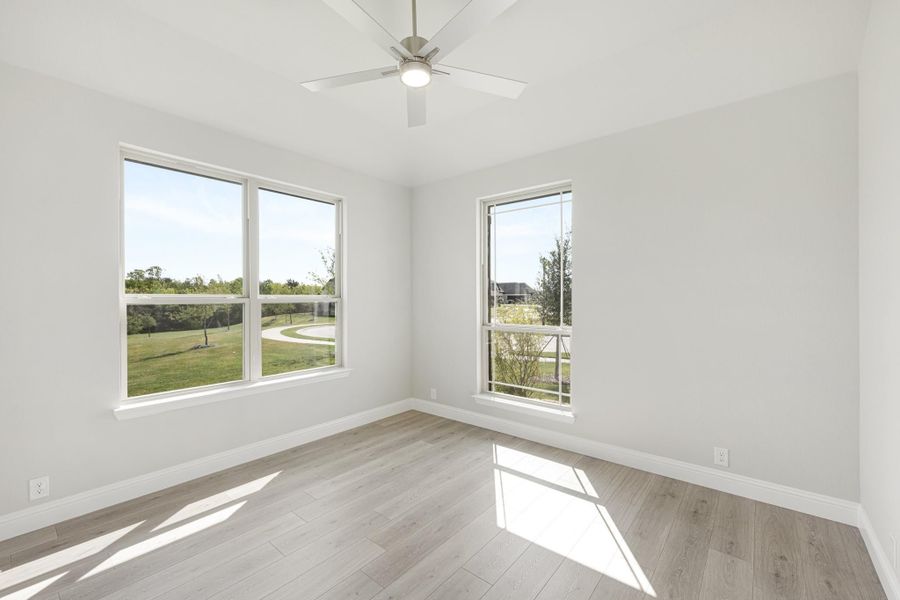 Empty room with light wood floors, ceiling fan, and two windows overlooking a green yard