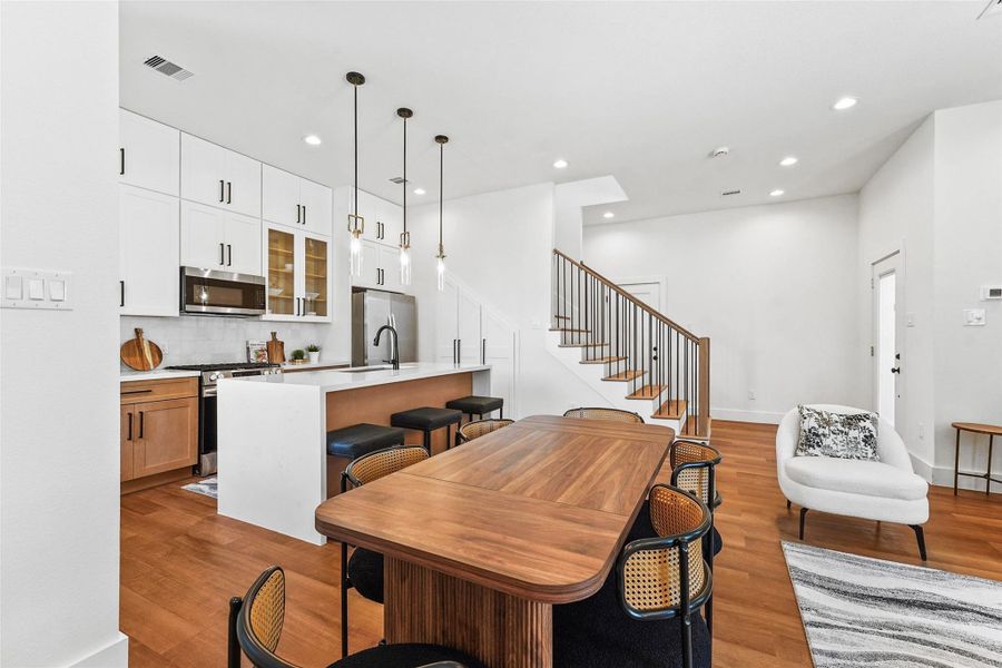 Modern kitchen with sleek white cabinetry, wood accents, quartz countertops, and a large island with pendant lighting. Open dining and living areas flow seamlessly, accented by a striking wrought iron staircase and warm wood flooring. Modern kitchen with sleek white cabinetry, wood accents, quartz countertops, and a large island with pendant lighting. Open dining and living areas flow seamlessly, accented by a striking wrought iron staircase and warm wood flooring.