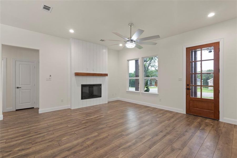 Spacious living area featuring wood-finish flooring, a contemporary fireplace with a white shiplap surround and wooden mantel, and a ceiling fan with integrated lighting Spacious living area featuring wood-finish flooring, a contemporary fireplace with a white shiplap surround and wooden mantel, and a ceiling fan with integrated lighting