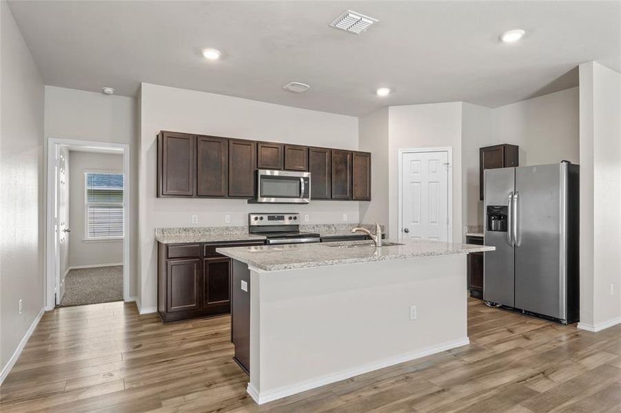 Kitchen featuring appliances with stainless steel finishes, dark brown cabinets, light wood-type flooring, an island with sink, and light stone countertops Kitchen featuring appliances with stainless steel finishes, dark brown cabinets, light wood-type flooring, an island with sink, and light stone countertops