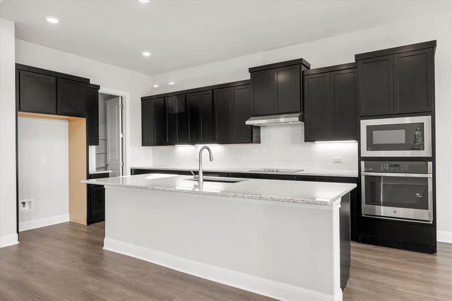 Kitchen featuring stainless steel appliances, an island with sink, dark cabinetry, light stone countertops, and dark wood-style floors