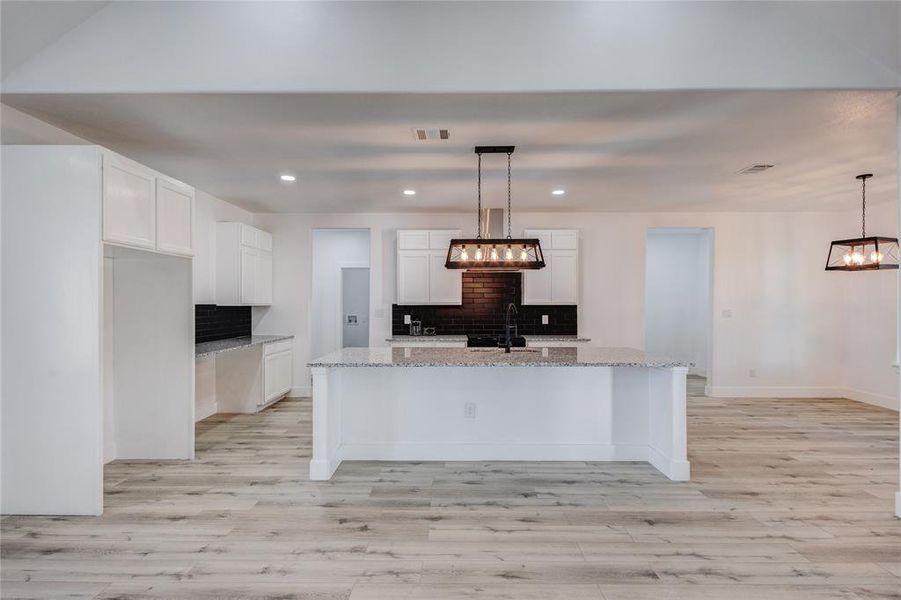 Kitchen with light stone counters, an island with sink, decorative light fixtures, backsplash, and white cabinetry