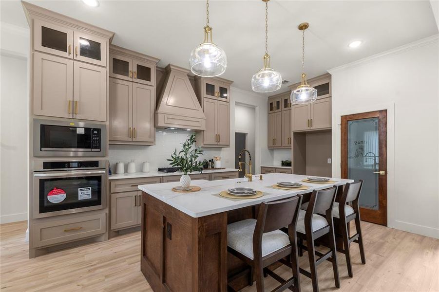 Kitchen featuring stainless steel appliances, custom range hood, light countertops, light wood-style flooring, and ornamental molding Kitchen featuring stainless steel appliances, custom range hood, light countertops, light wood-style flooring, and ornamental molding