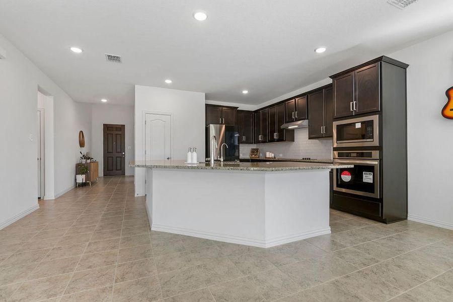 Kitchen featuring dark wood finish cabinets, light stone counters, stainless steel appliances, an island with sink, and recessed lighting Kitchen featuring dark wood finish cabinets, light stone counters, stainless steel appliances, an island with sink, and recessed lighting