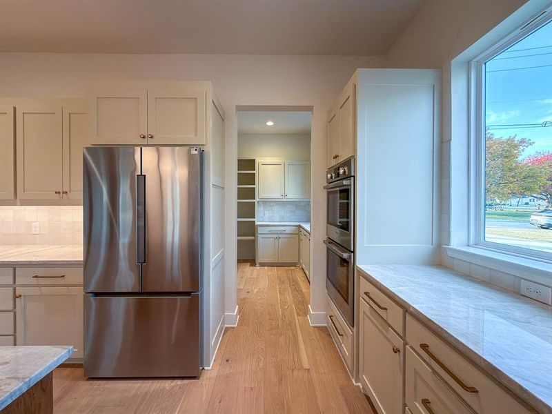Kitchen featuring stainless steel appliances, light wood-finish flooring, white cabinetry with gold-tone hardware, a white tile backsplash, and light-toned countertops