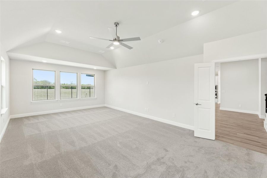 Empty living room with vaulted ceiling, ceiling fan, gray carpet, and triple windows overlooking a patio.