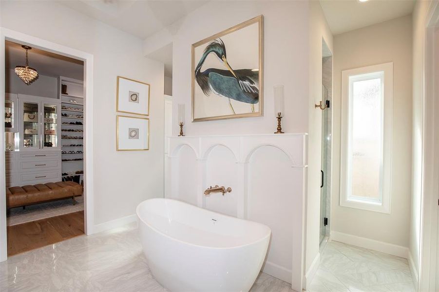 This bathroom features a modern freestanding tub, elegant white paneling, and a large bird artwork. It connects to a spacious walk-in closet with ample shelving and a chandelier, offering a luxurious and functional space.