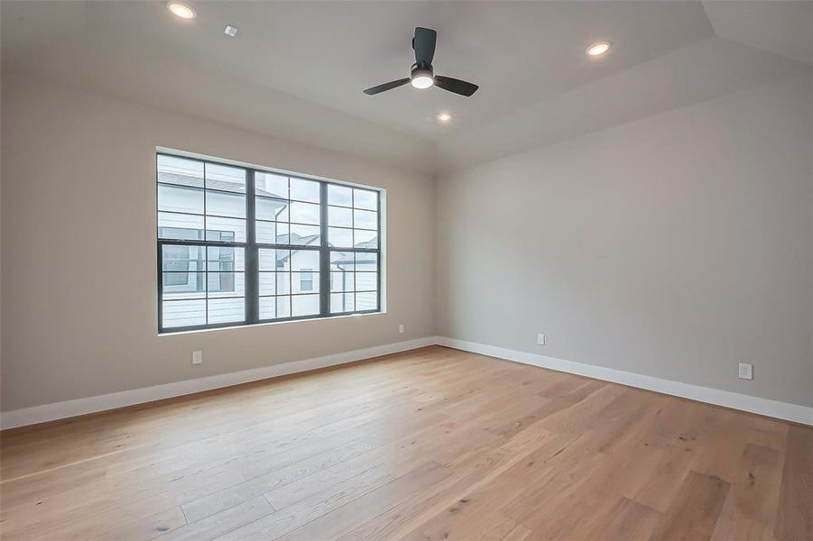 Bright and spacious room with light wood floors, neutral walls, and a large window for abundant natural light. Features recessed lighting and a ceiling fan for a modern, inviting feel.