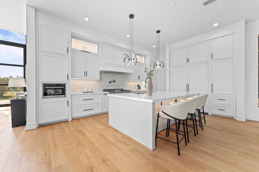 This kitchen features a modern, open design with sleek white cabinetry, a large island with seating, and contemporary pendant lighting. It boasts high-end appliances, ample storage, and light wood flooring, offering a bright and inviting atmosphere.
