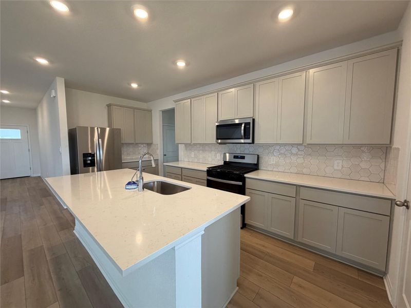 Kitchen featuring light stone countertops, stainless steel appliances, an island with sink, light wood-style flooring, and gray cabinets