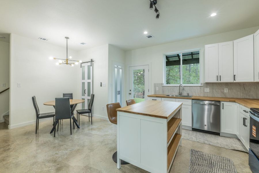 Kitchen and dining area featuring concrete flooring, a central island with wood-finish countertop, white cabinetry with matte black hardware, stainless steel appliances, and a modern chandelier Kitchen and dining area featuring concrete flooring, a central island with wood-finish countertop, white cabinetry with matte black hardware, stainless steel appliances, and a modern chandelier