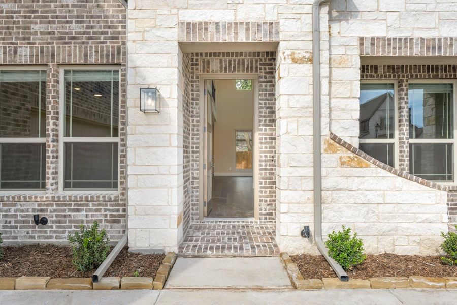 Home entrance with brick and stone exterior, open front door, wall lantern, and small landscaped shrubs.