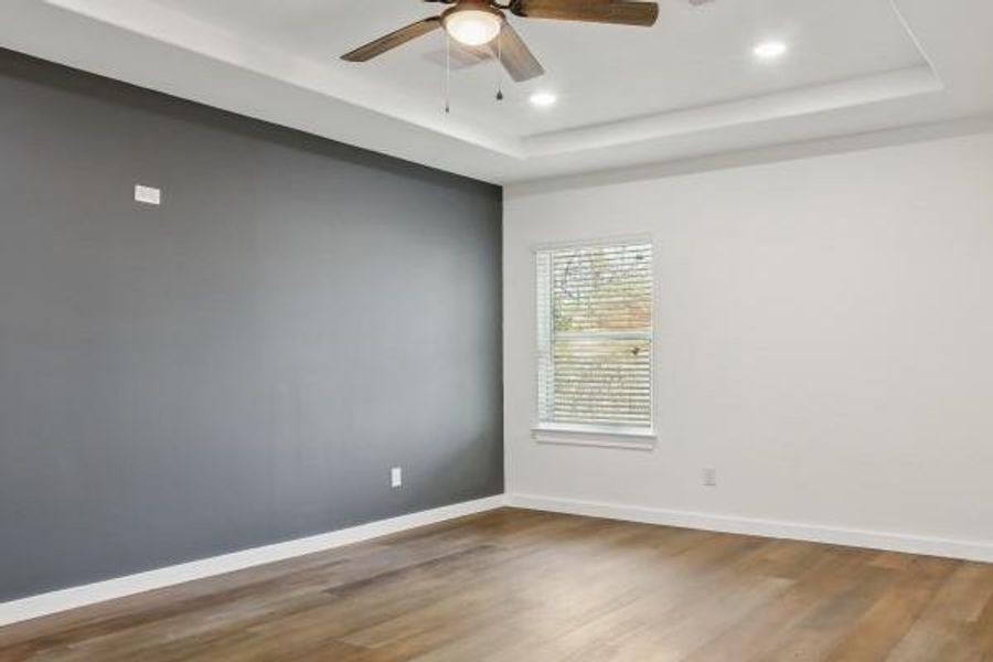 Spacious primary bedroom featuring a stylish accent wall, elegant tray ceiling with recessed lighting, and warm vinyl plank flooring. Spacious primary bedroom featuring a stylish accent wall, elegant tray ceiling with recessed lighting, and warm vinyl plank flooring.