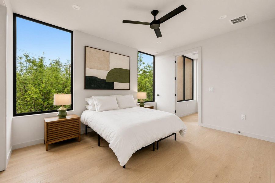 Tall windows frame verdant outdoor views in this naturally illuminated bedroom with light-toned hardwood flooring.