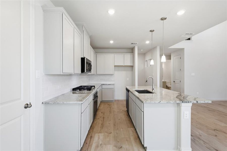 Kitchen featuring decorative backsplash, light stone countertops, light wood-type flooring, hanging light fixtures, and a center island with sink