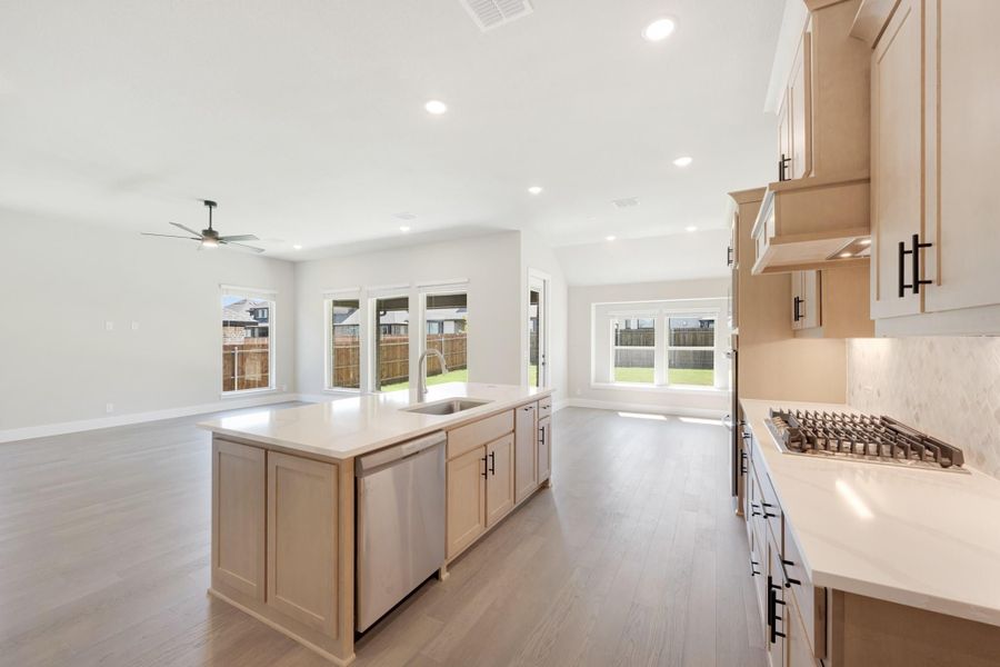 Open kitchen with light wood cabinets, center island with sink, and gas cooktop open to living area with hardwood floors