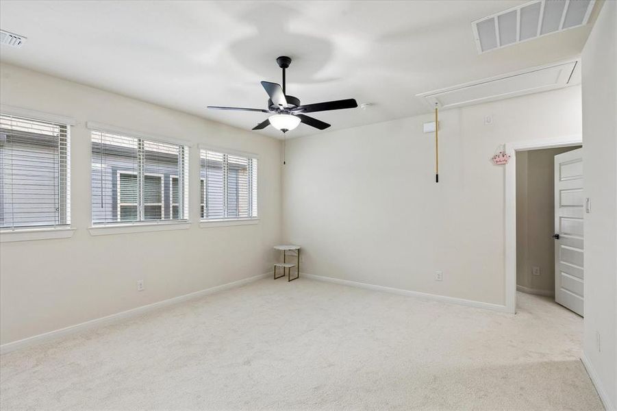Expansive carpeted room featuring a ceiling fan with integrated lighting, three windows with blinds, a white paneled door, and an overhead attic access panel Expansive carpeted room featuring a ceiling fan with integrated lighting, three windows with blinds, a white paneled door, and an overhead attic access panel