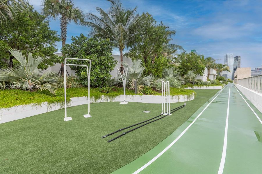 Outdoor fitness area featuring a synthetic running track, multi-use exercise frames, manicured landscaping with mature palms, and tiered retaining walls