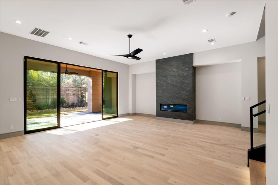 This spacious living room features large sliding glass doors opening to a backyard, a sleek modern fireplace, light wood flooring, and a ceiling fan, offering a bright and airy atmosphere.