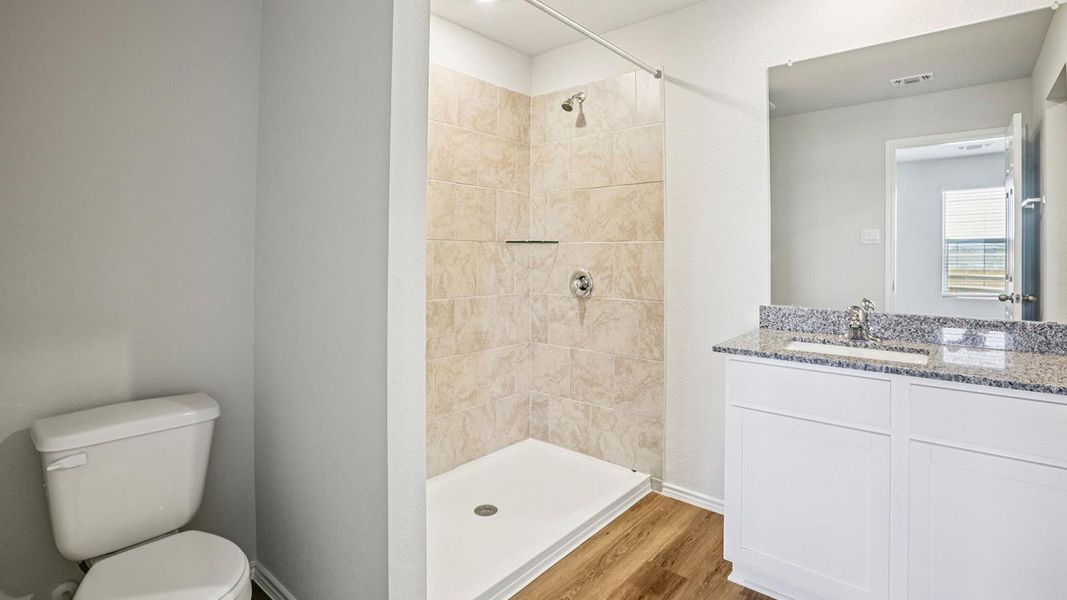 Bathroom featuring a tiled standing shower with chrome fixtures, a single vanity with a granite countertop and undermount sink, a large wall-mounted mirror, and wood-finish flooring Bathroom featuring a tiled standing shower with chrome fixtures, a single vanity with a granite countertop and undermount sink, a large wall-mounted mirror, and wood-finish flooring