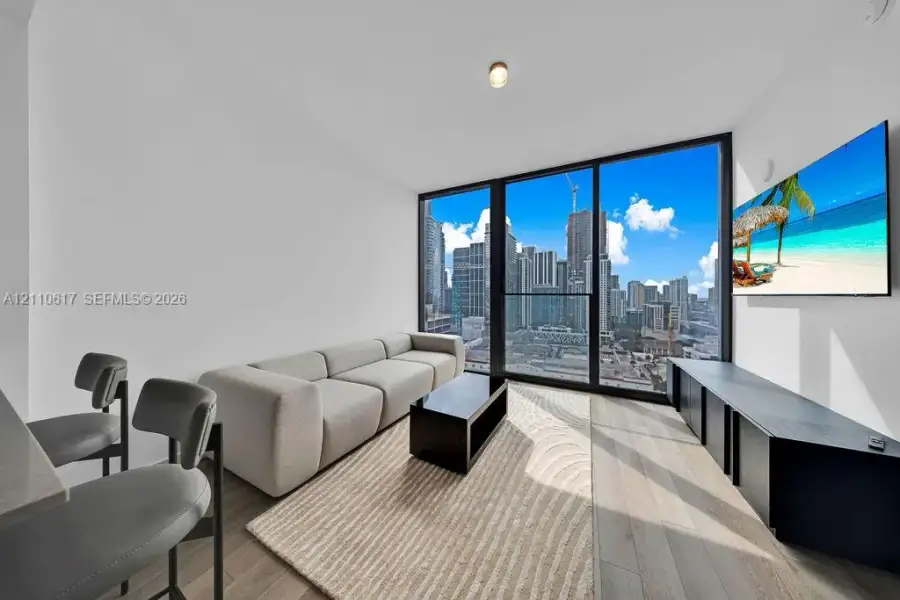Floor-to-ceiling windows featuring urban skyline scenery, wood-finish flooring, and a sleek matte black built-in media console Floor-to-ceiling windows featuring urban skyline scenery, wood-finish flooring, and a sleek matte black built-in media console
