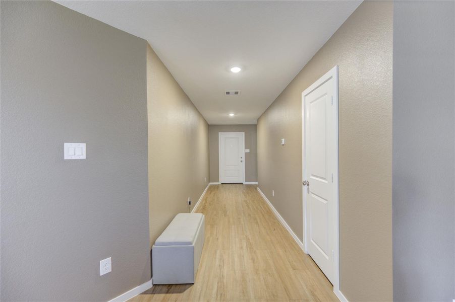 This photo shows a well-lit, narrow hallway with light vinyl plank flooring and neutral  walls. It features recessed lighting, a white interior door, and a small bench, offering a clean and modern entryway feel.