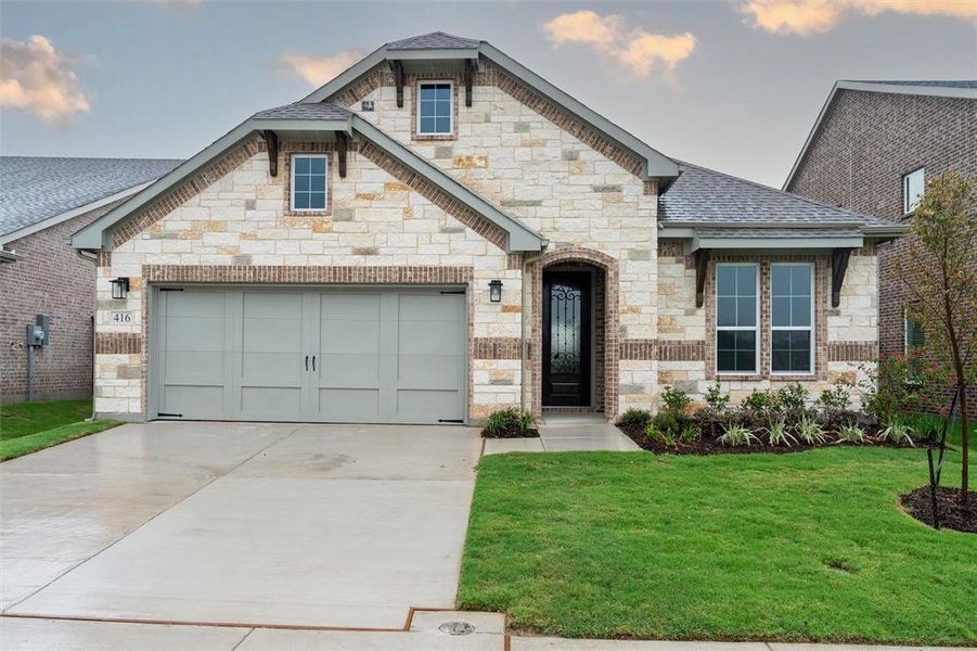 French country home featuring a front yard, an attached garage, concrete driveway, stone siding, and brick siding