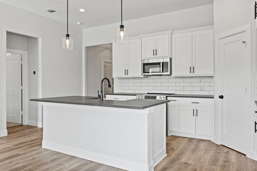 Kitchen featuring white cabinets, tasteful backsplash, stainless steel microwave, an island with sink, and hanging light fixtures