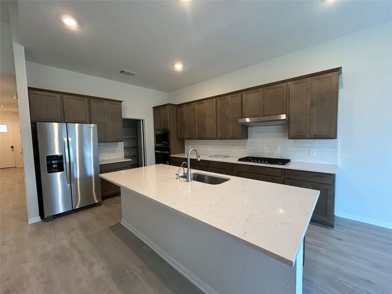 Kitchen with decorative backsplash, dark brown cabinets, appliances with stainless steel finishes, light stone counters, and an island with sink Kitchen with decorative backsplash, dark brown cabinets, appliances with stainless steel finishes, light stone counters, and an island with sink