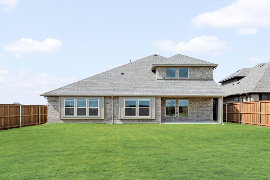 Rear view of brick home with large windows, covered patio, and fenced backyard with green lawn