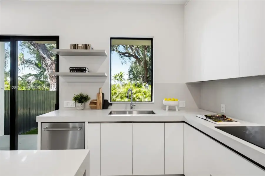 Minimalist kitchen featuring matte white flat-panel cabinetry, light-toned stone-surface countertops, stainless steel undermount sink, built-in dishwasher, and floating wall shelves